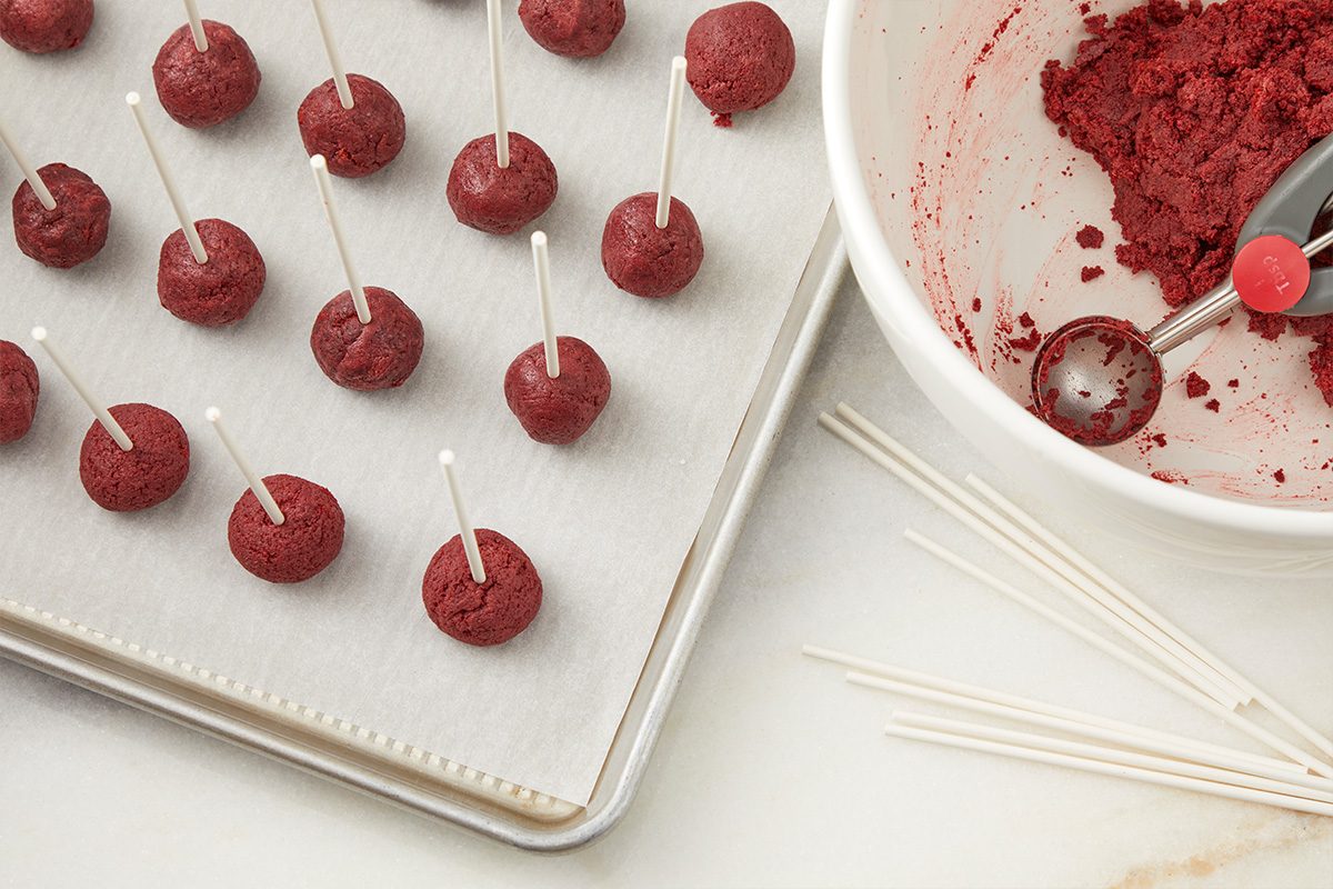 Round red cake pops with sticks are arranged on a parchment-lined baking sheet. Nearby, a white bowl contains red cake mixture, a scoop, and some extra sticks on the counter.