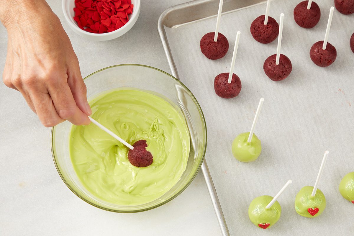 A hand dips a cake pop into a bowl of green icing. Nearby, undipped and dipped cake pops are on a tray, some with small red heart decorations. A bowl of red sprinkles sits in the background.
