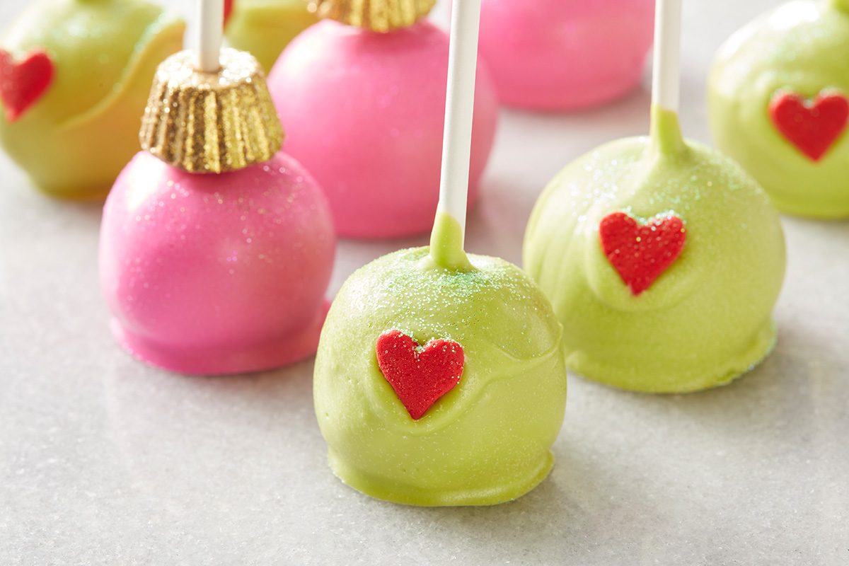 Cake pops decorated in green and pink, with red heart shapes on the side. Some have gold wrappers on top, resembling Christmas ornaments, all arranged on a light surface.