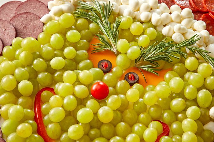 Closeup shot of a festive platter arranged to look like the Grinch, made with green grapes, mozzarella balls, rosemary, a cherry tomato, red pepper, yellow cheese, and black olives for facial features