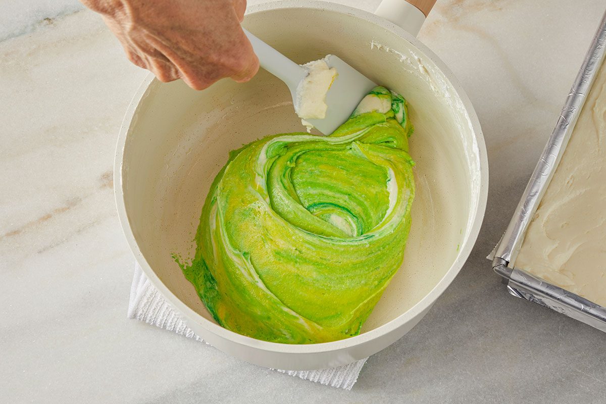 Overhead shot of a hand uses a spatula to swirl green food coloring into cream cheese mixture in a white bowl; sitting on a marble countertop next to a rectangular pan lined with parchment paper