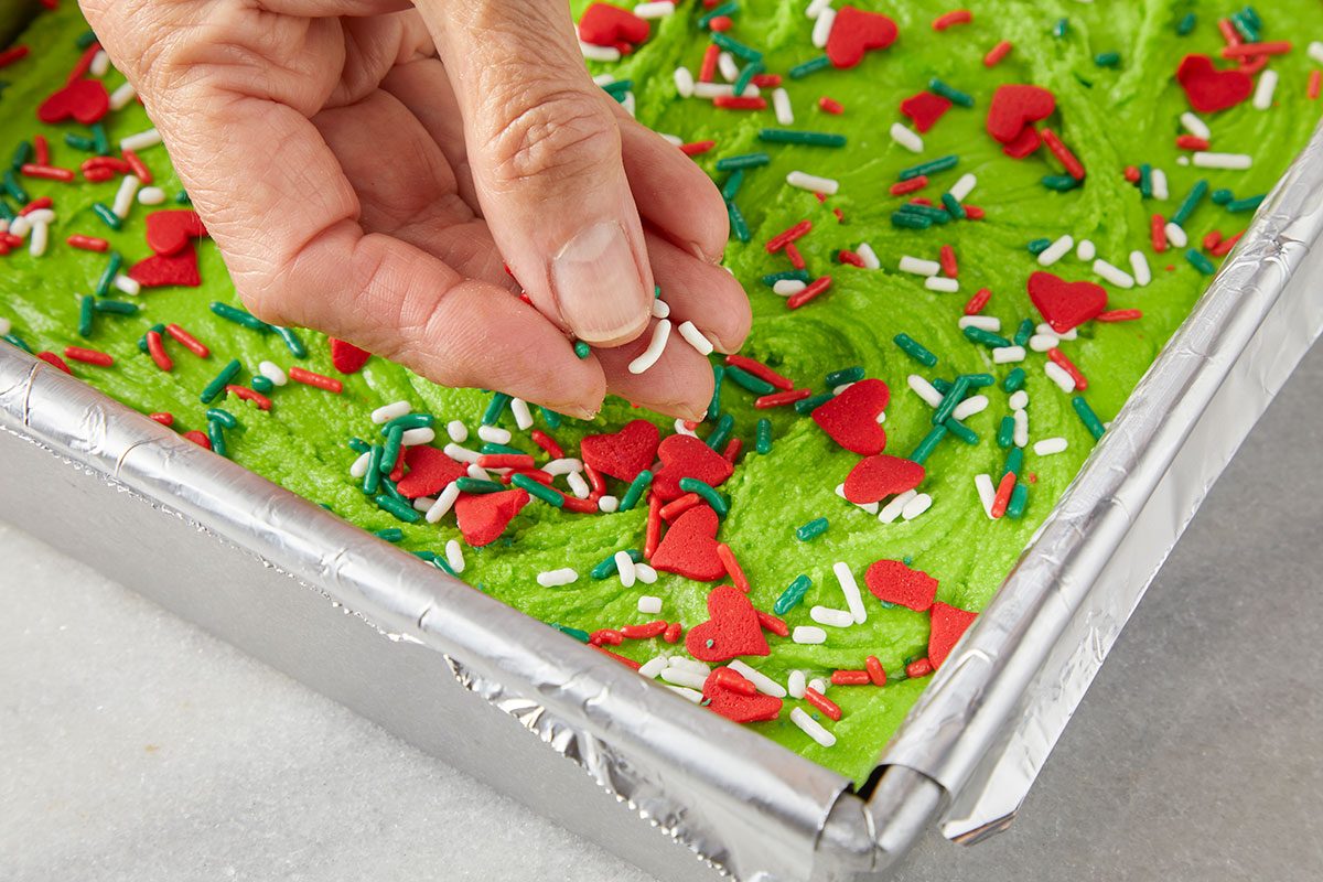 3/4th shot of a hand sprinkles colorful heart shaped and rod shaped sprinkles onto green batter in a foil lined baking pan