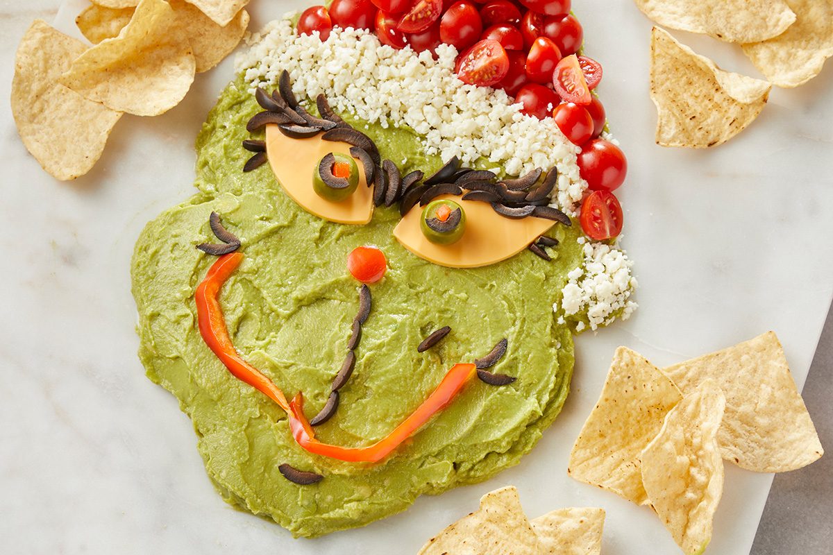 Overhead shot of a dip platter arranged as the Grinch’s face; made from guacamole; cheese; tomatoes; sliced olives; chips; and vegetables with tortilla chips placed around the platter