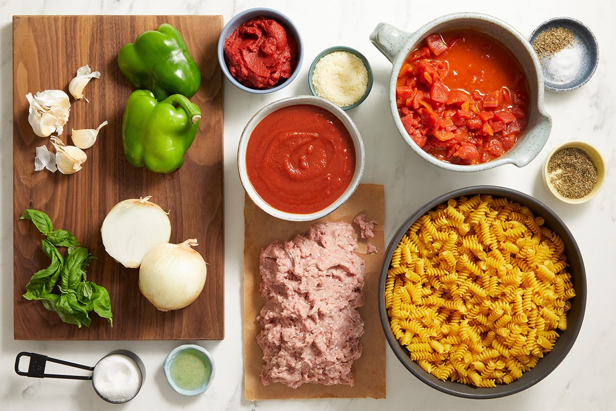 Chopped green bell peppers, garlic, onion, and basil on a cutting board; uncooked ground meat, grated cheese, seasonings, canned tomatoes, tomato sauce, and cooked macaroni pasta arranged on a counter.