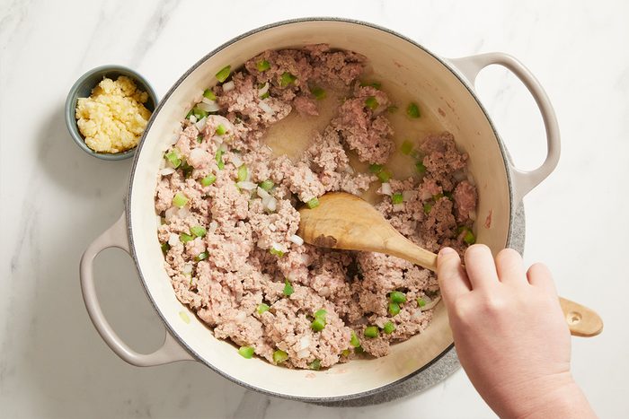 A hand stirs ground meat, chopped onions, and green bell peppers in a large pot with a wooden spoon. A small bowl of minced garlic sits nearby on a white marble surface.