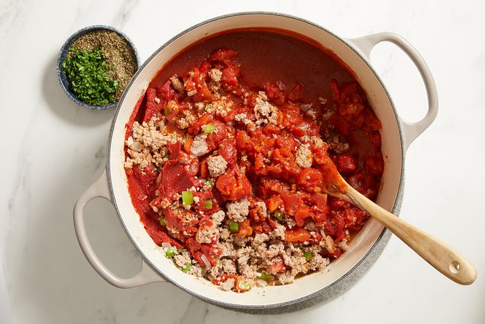 A large white pot filled with chili, containing ground meat, beans, tomatoes, and chopped vegetables, sits on a white surface. A wooden spoon rests in the pot, and a small bowl of chopped herbs and spices is nearby.