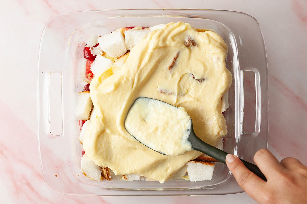 A hand spreads creamy yellow pudding over chunks of white cake and red fruit in a glass baking dish on a light pink surface.