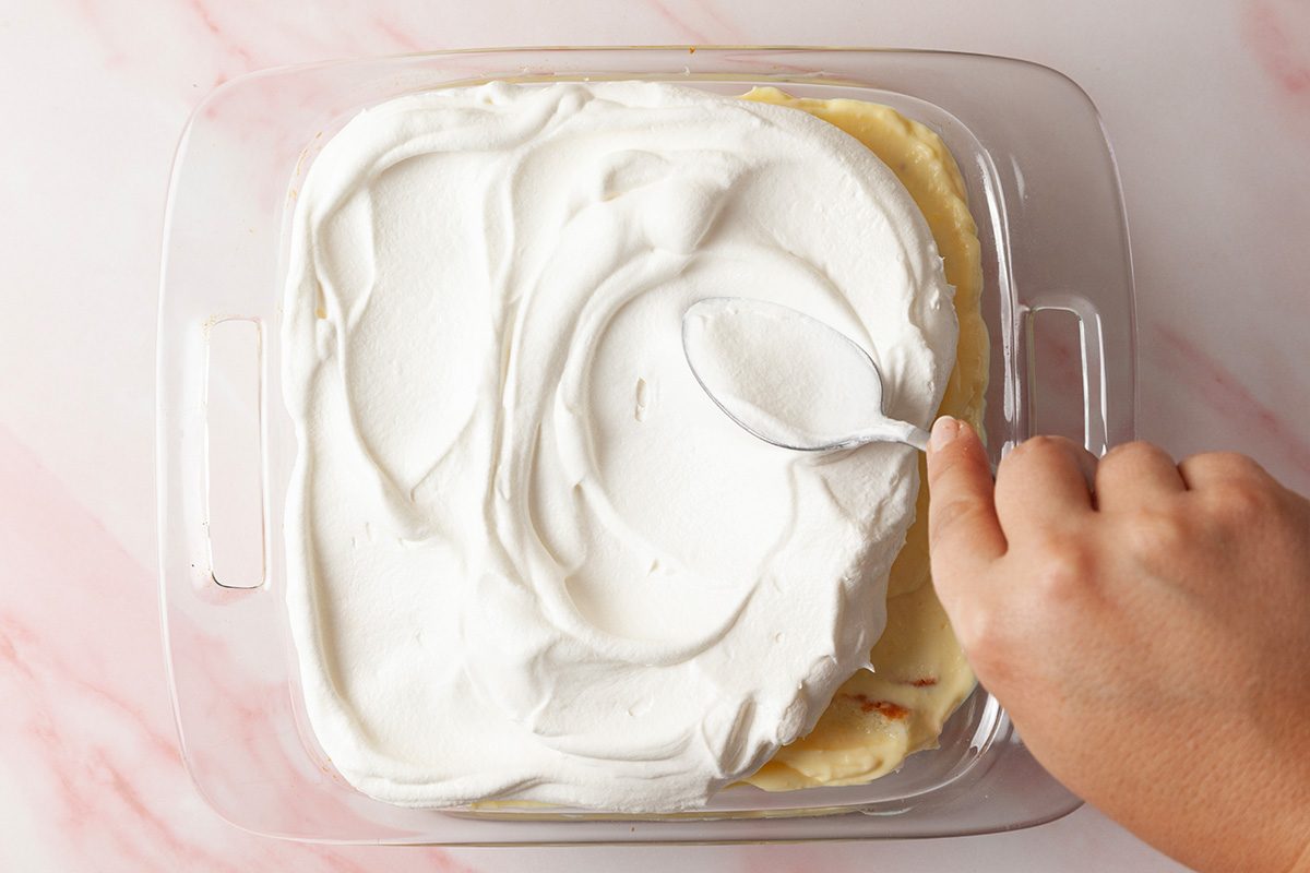 A hand uses a spoon to spread whipped cream over a layered dessert in a square glass dish on a light pink marble surface.