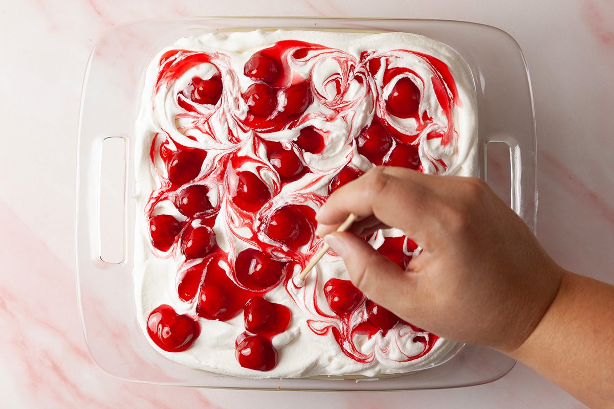 A hand swirls cherry topping into a layer of whipped cream in a glass baking dish, creating a marbled effect on a dessert.