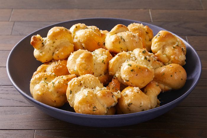 A blue bowl filled with golden-brown garlic knots topped with herbs and grated cheese, placed on a wooden table.