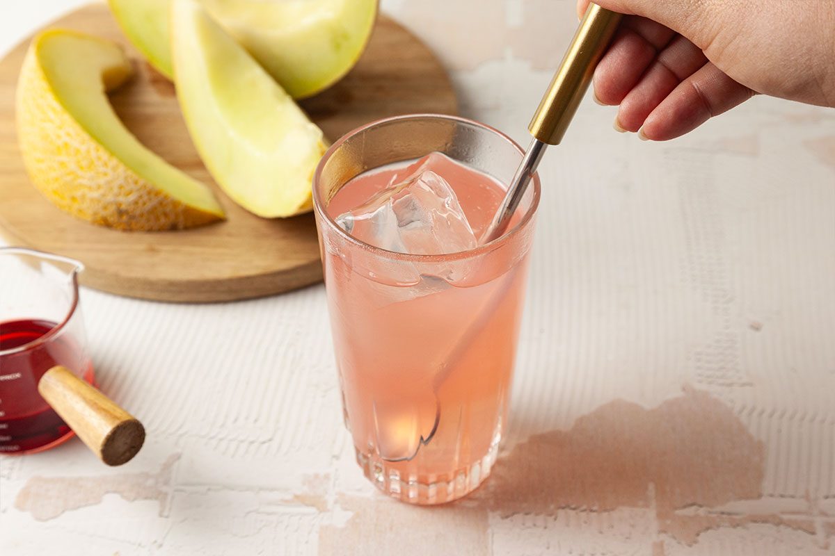 A hand stirs a pink drink with ice in a glass, using a metal stirrer. In the background, there are sliced pieces of melon on a wooden board and a small glass container with red liquid.