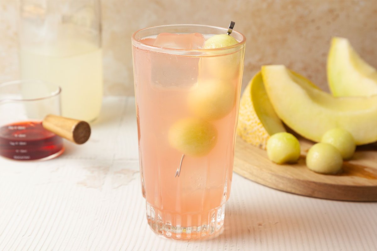A tall glass of pink lemonade with ice and two melon balls on a skewer, set on a white surface. In the background are melon slices, round melon balls, a small jar of red liquid, and a bottle of lemonade.