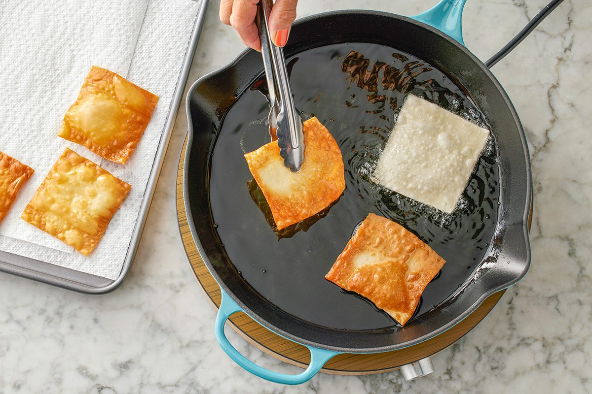 This is a close-up shot; a hand uses tongs to flip a wonton wrapper frying in hot oil; golden cooked wontons rest on a paper towel-lined tray on a marble countertop