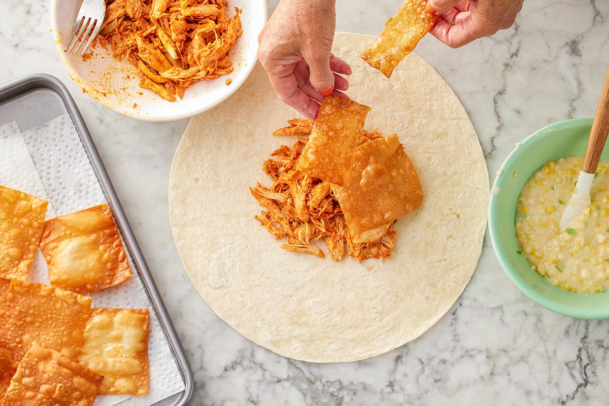 This is an overhead shot showing hands assembling a wrap with shredded chicken and crispy wonton chips on a tortilla; background includes bowls of chicken; egg wash; extra chips on a tray atop a marble surface