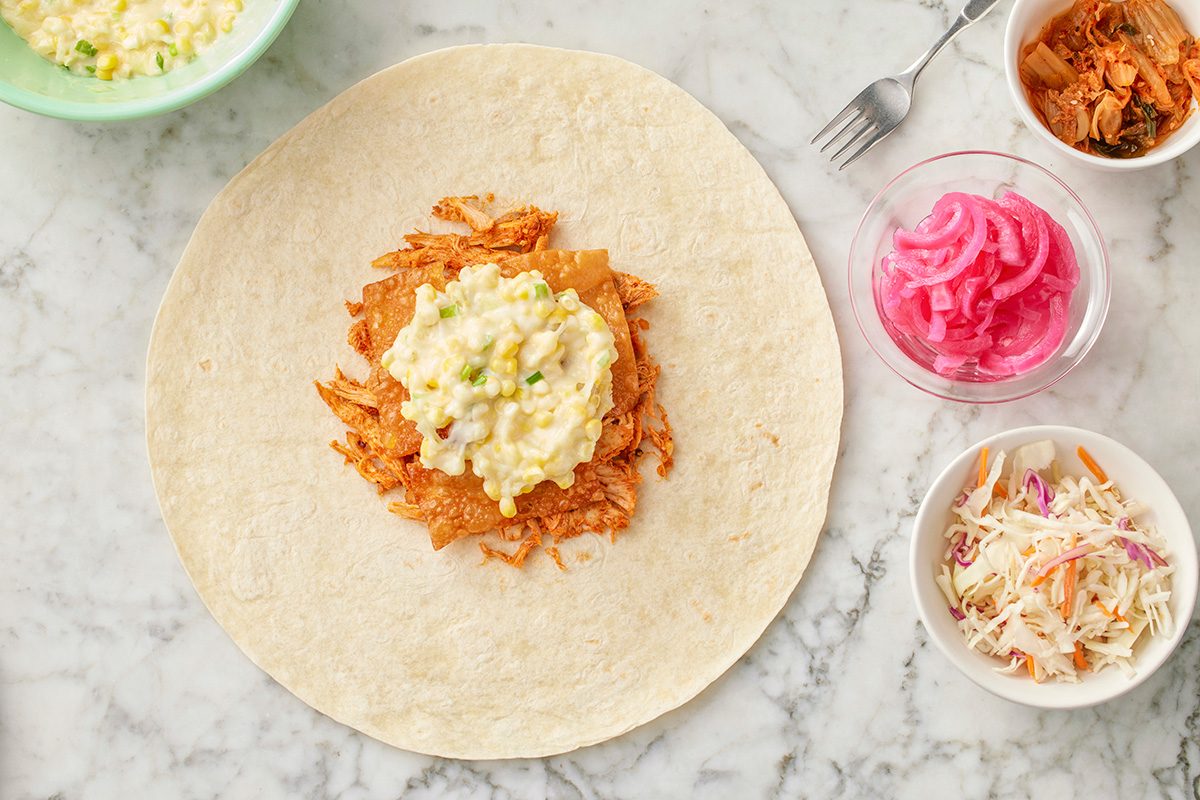 This is an overhead shot; a flour tortilla on marble holds shredded meat; corn salad; cheese; surrounded by bowls of coleslaw; pickled onions; extra fillings; A fork lies beside everything, highlighting a fresh meal setting