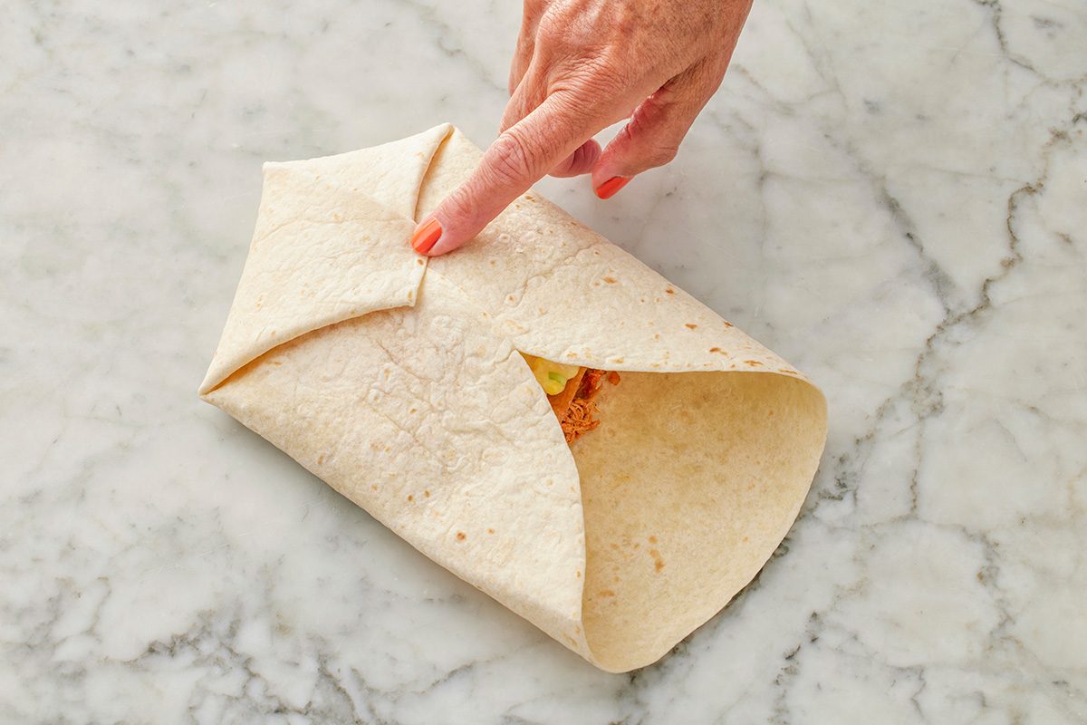 This top-down shot shows a hand folding a tortilla wrap with assorted ingredients on a marble surface; instructively demonstrating proper wrapping technique