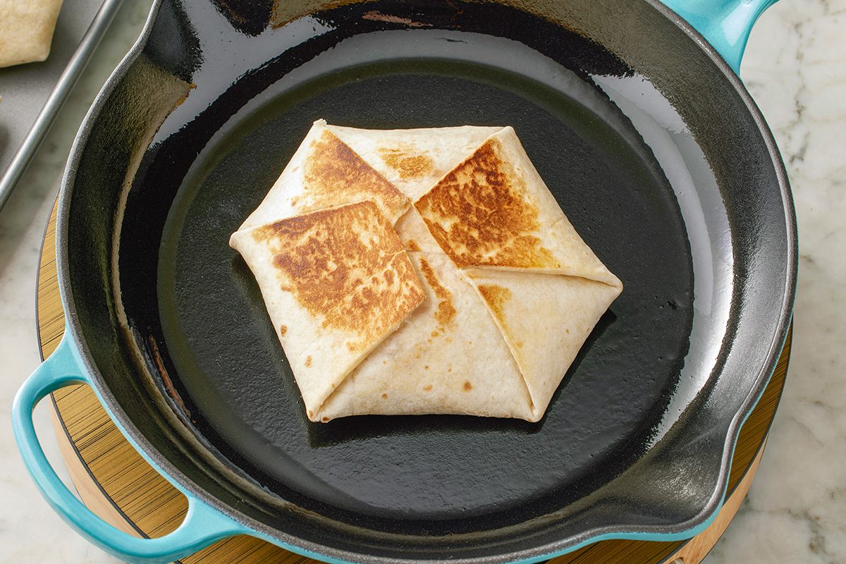 This is a close-up food shot; a folded and grilled tortilla wrap rests in a blue-handled cast iron skillet on a stovetop; another wrap is visible on a metal tray in the background