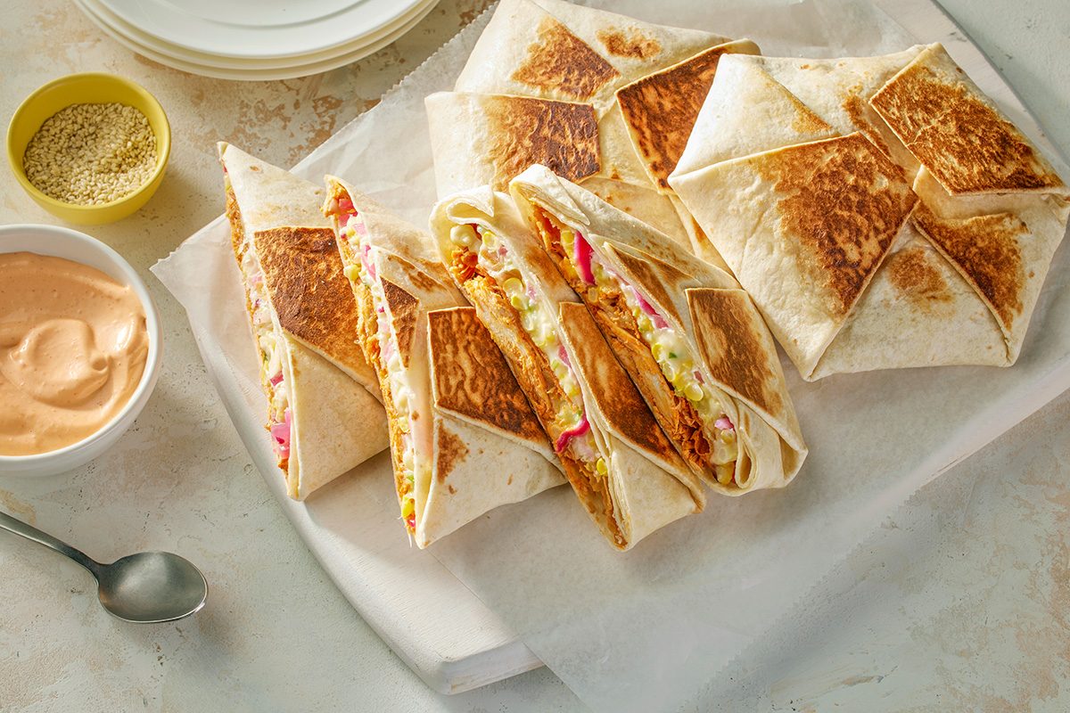 This is a close-up shot of folded grilled tortilla wraps stuffed with vibrant vegetables and chicken on a white board; accompanied by creamy dipping sauce; sesame seeds; garnished stylishly against a rustic background