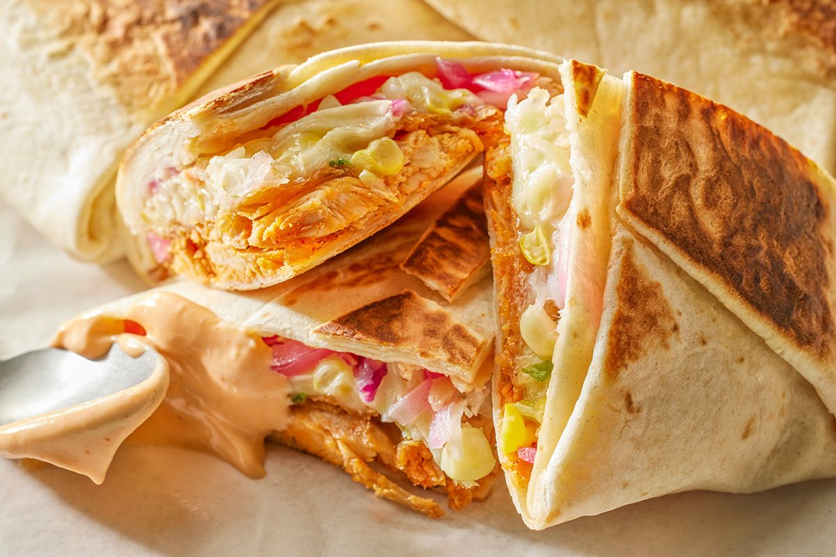 This is a close-up shot of a grilled burrito cut in half; exposing juicy chicken; melted cheese; and colorful vegetables inside; with a creamy dipping sauce on the side against a neutral background