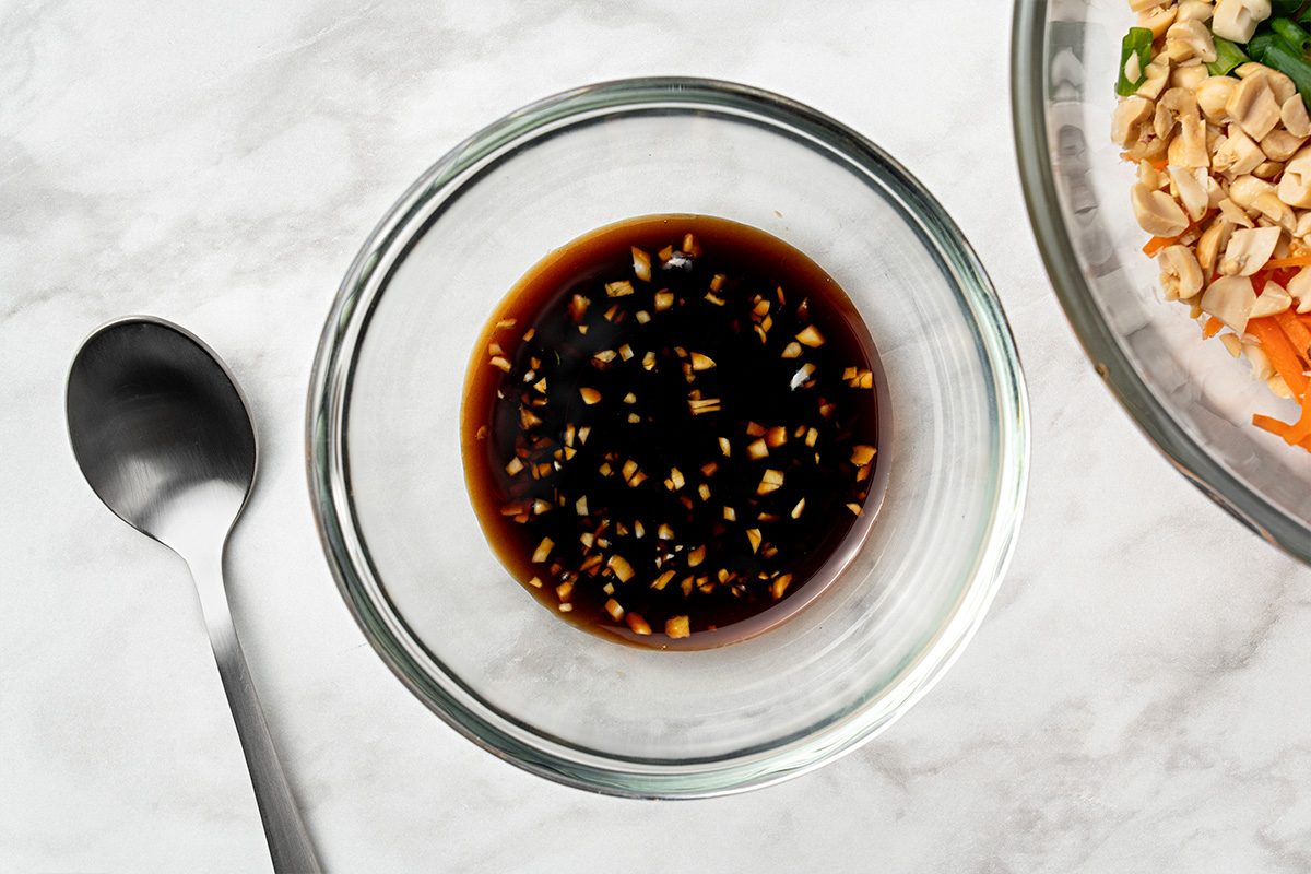 A glass bowl filled with a dark soy-based sauce containing minced garlic or ginger sits on a marble surface next to a spoon and a partial bowl of chopped vegetables and nuts.