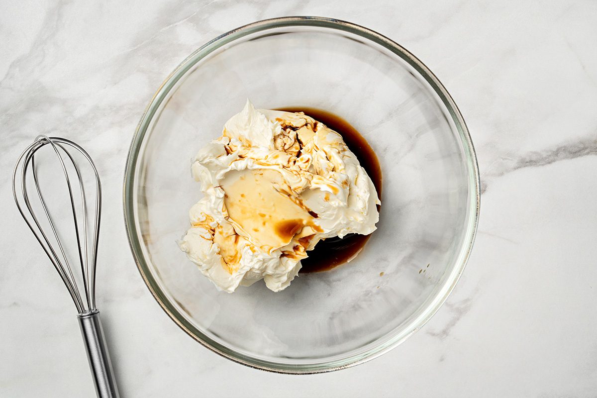 A glass bowl with cream cheese, vanilla extract, and sugar on a white marble surface, next to a metal whisk.