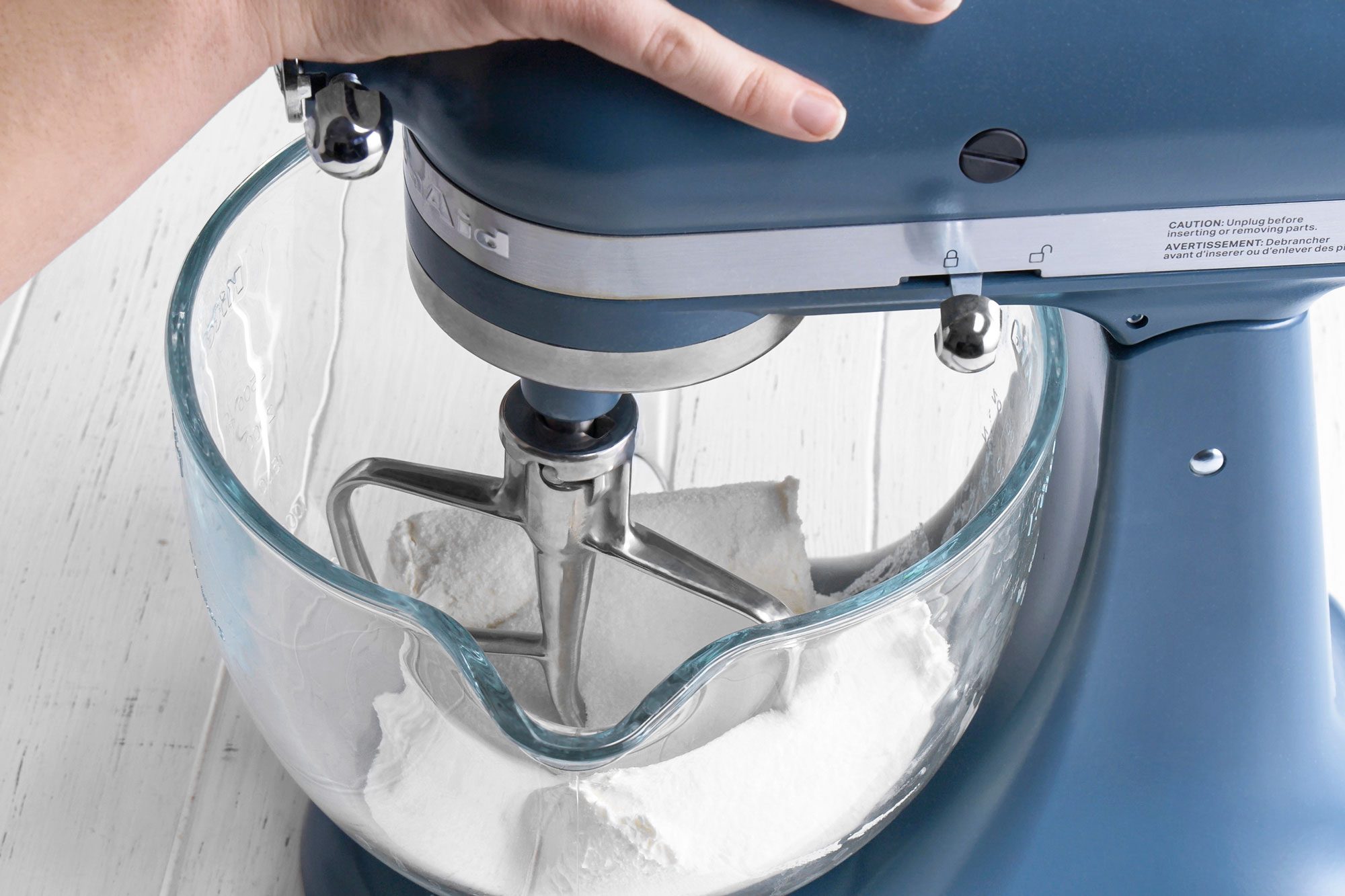 3/4th shot of a hand operates a blue stand mixer with a glass bowl containing white powder, likely flour or sugar, on a white wooden surface
