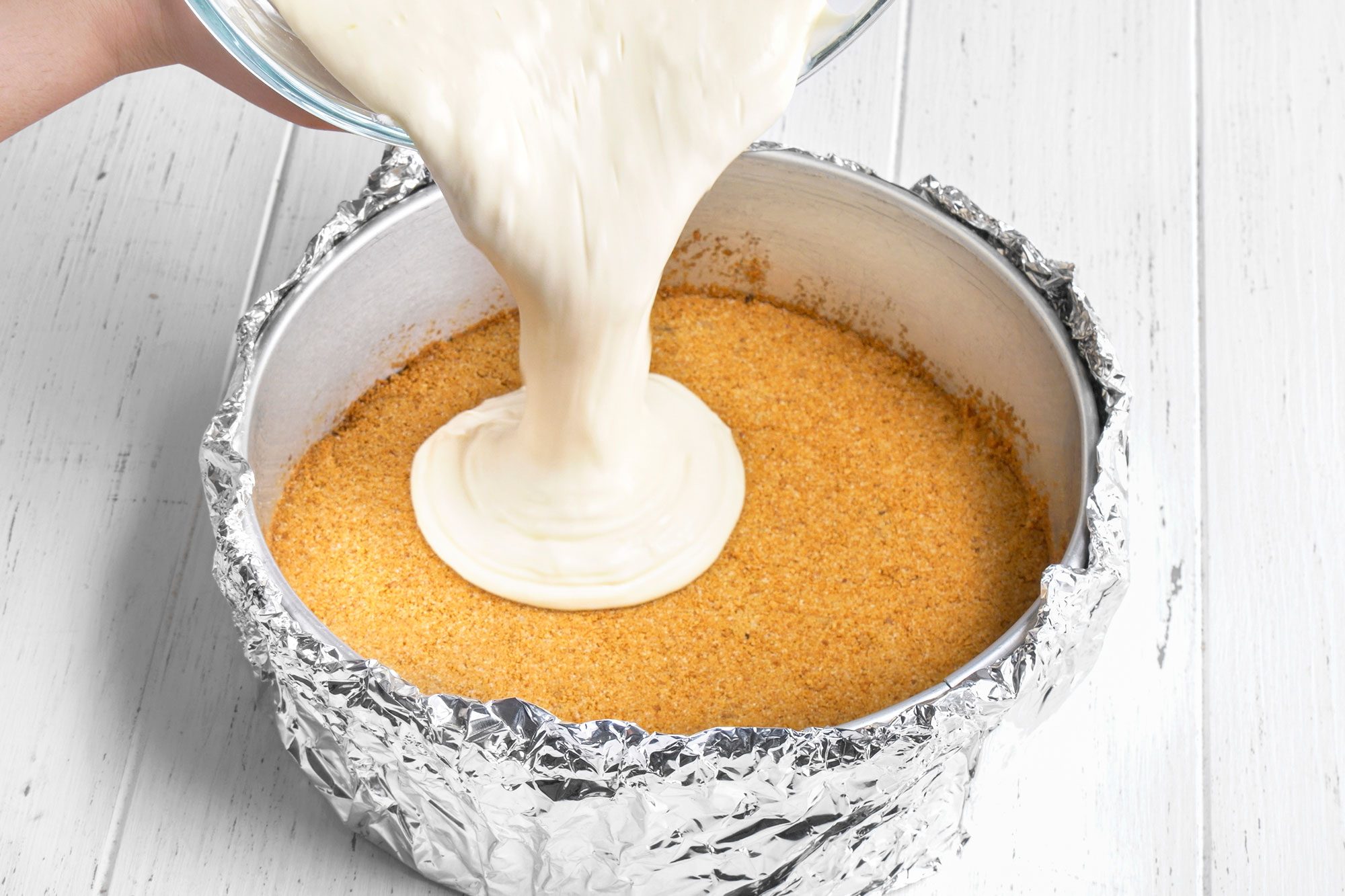 3/4th shot of a hand pours creamy cheesecake filling over a crumbly graham cracker crust in a foil wrapped round baking pan on a white wooden surface