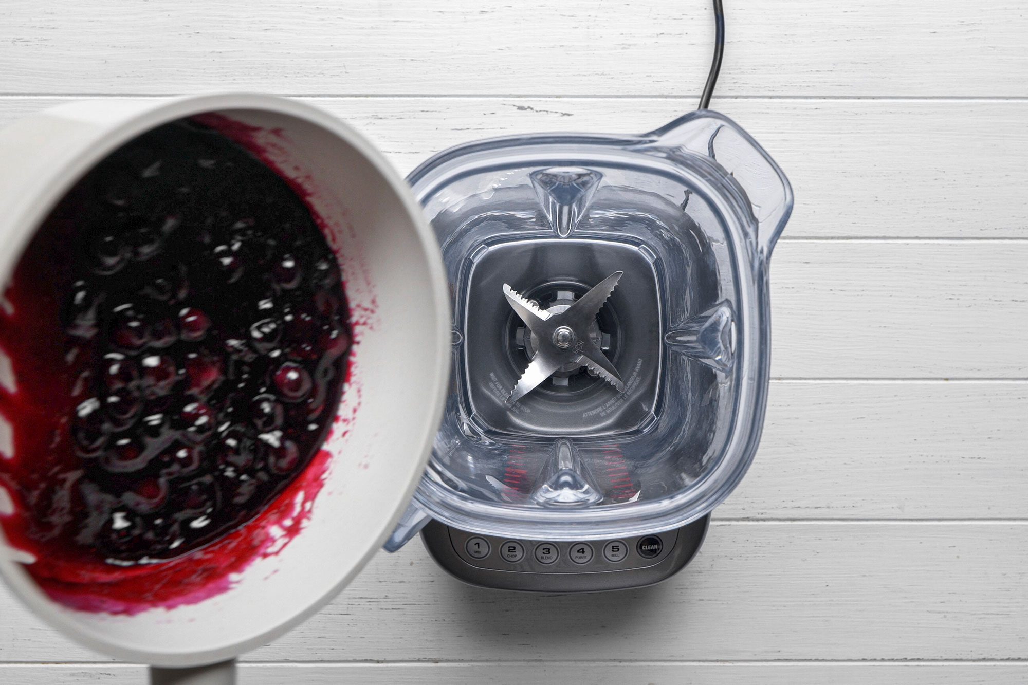 overhead shot of a clear blender sits on a white wooden surface, viewed from above, Next to it, a white pan containing a dark, berry colored mixture is being tilted toward the blender