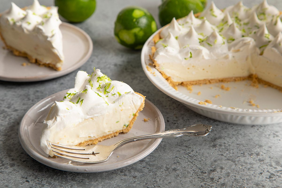 A creamy key lime pie topped with whipped cream sits on a plate with a fork, while the rest of the pie and whole limes are in the background.