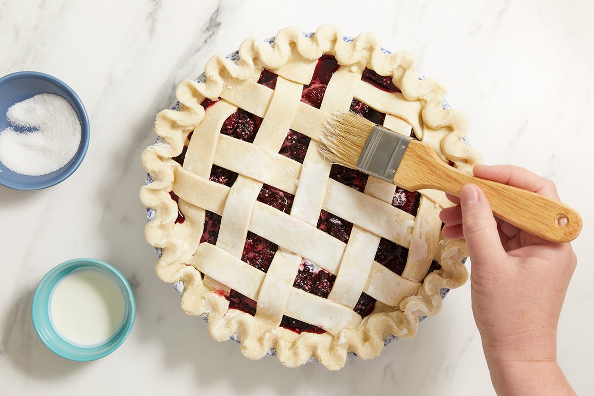 A hand brushes milk over a lattice pie crust, with small bowls of sugar and milk nearby on a white marble surface.