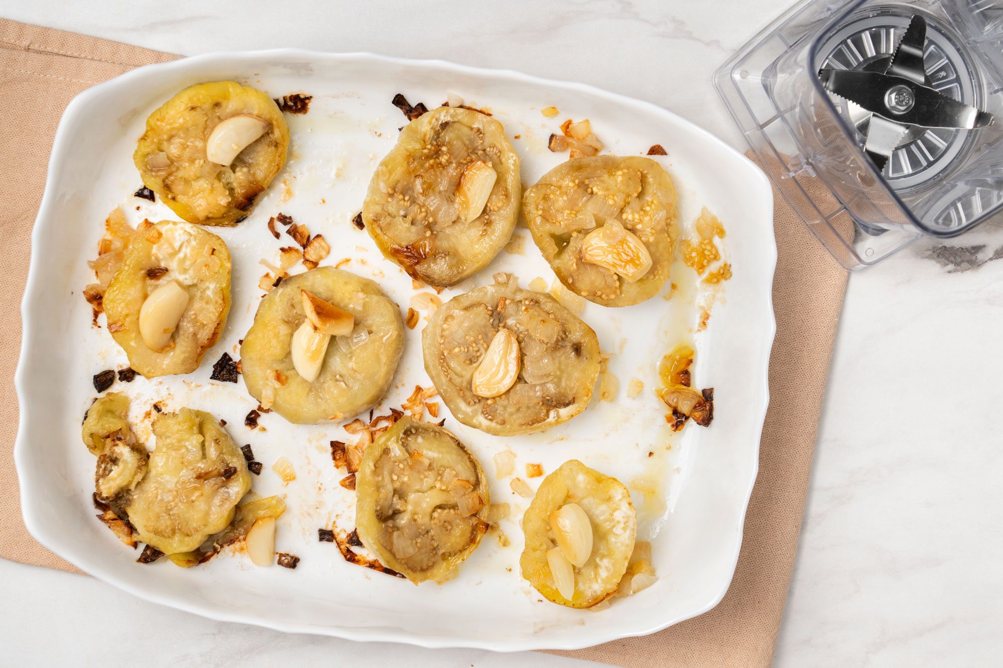 Overhead shot of a white baking dish holds roasted round yellow squash slices topped with sautéed onions and garlic cloves, sitting on a tan placemat near a food processor on a marble surface;