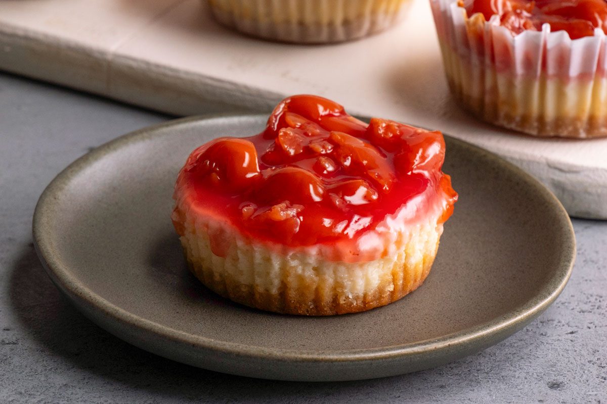 3/4th shot of a mini cherry cheesecake topped with glossy cherry pie filling sits on a gray plate, with more cheesecakes in paper liners visible in the background on a light serving board