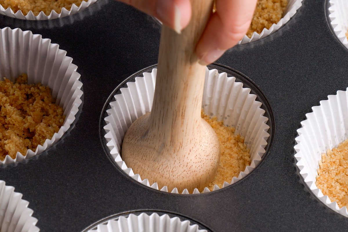 3/4th shot of a hand presses crushed graham cracker crumbs into paper cupcake liners in a muffin pan using a wooden tamper, preparing crusts for desserts