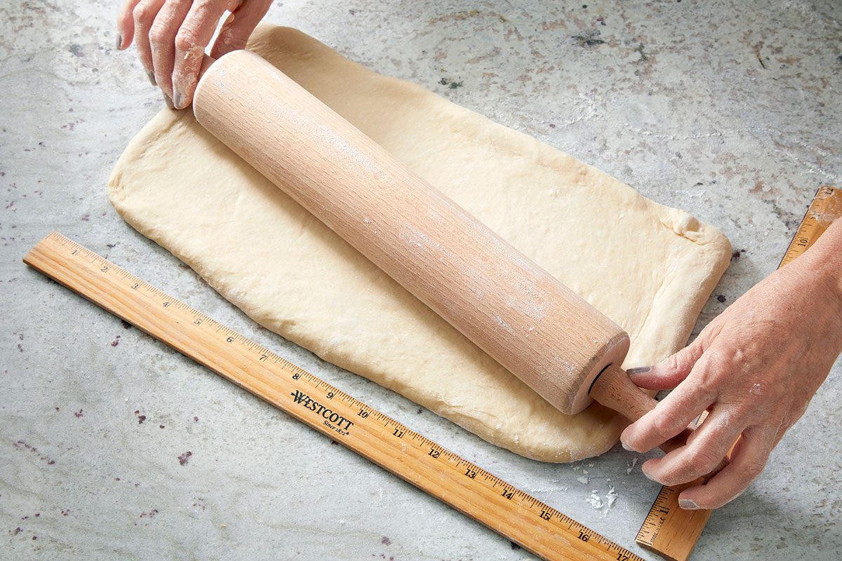 Overhead shot of hands rolling out dough with a wooden rolling pin on a floured surface; with a wooden ruler placed alongside for measurement;