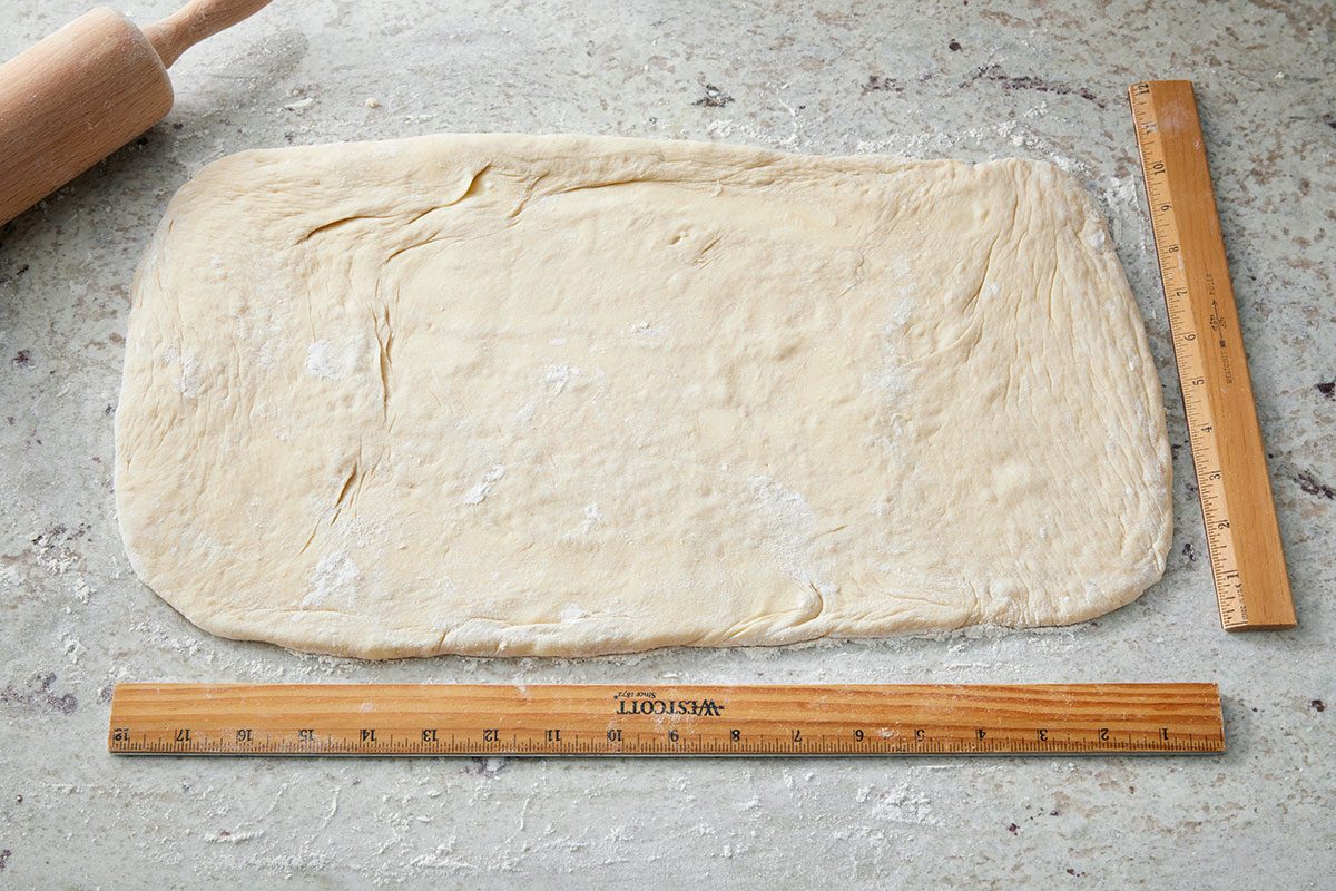 High-angle shot of a rectangular sheet of dough rolled out on a floured surface; with a wooden rolling pin placed at the upper left and two wooden rulers measuring its length and width.