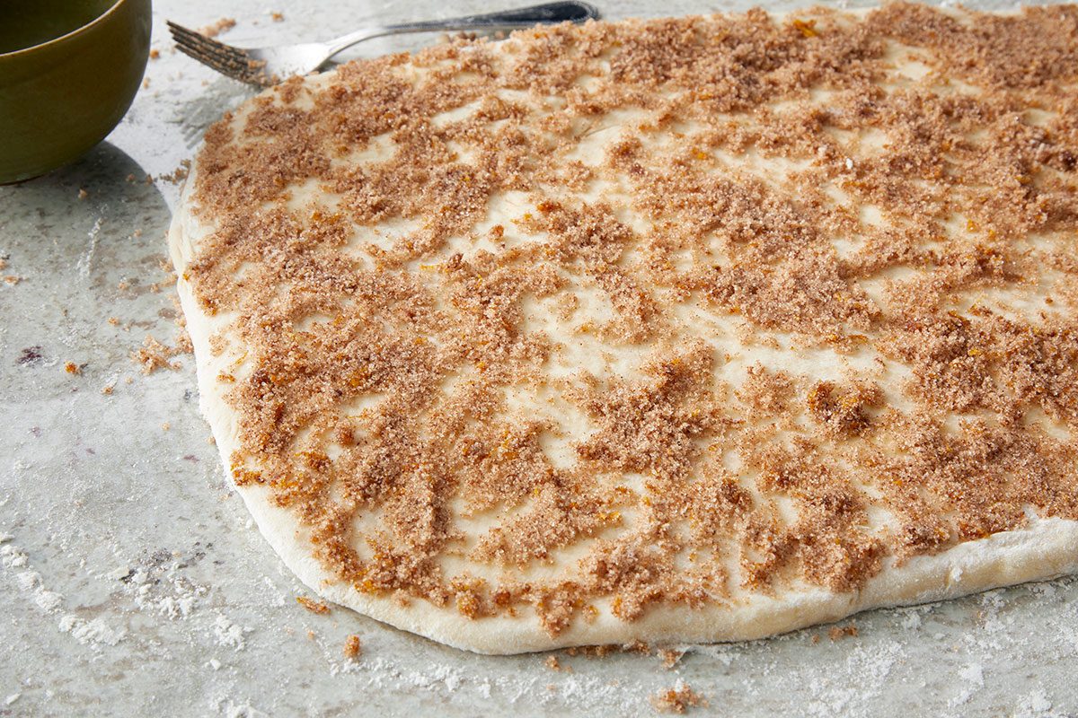 Close-up shot of a sheet of raw dough spread on a floured surface; sprinkled with brown sugar and cinnamon; ready to be rolled; a fork and part of a green bowl are visible in the background.