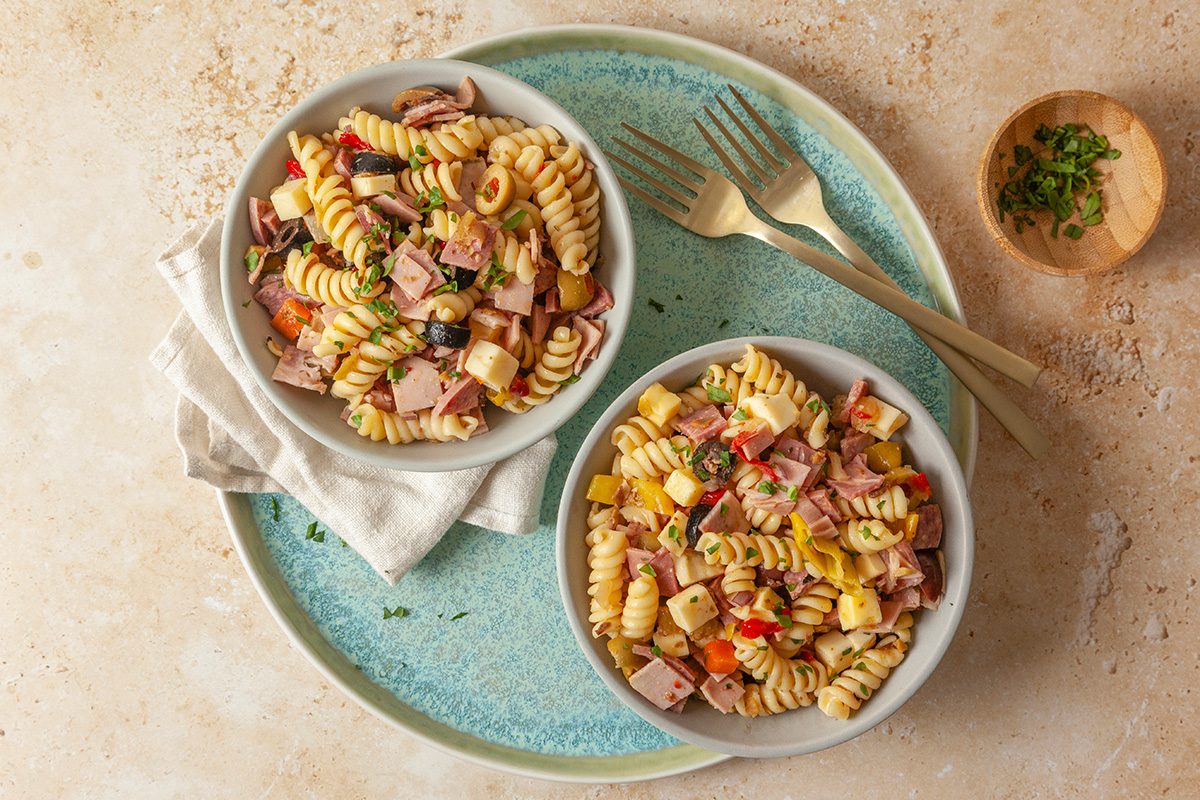 overhead shot of Two bowls of pasta salad with rotini, diced ham, cheese, olives, and peppers sit on a blue plate; Gold forks and a small bowl of chopped herbs are nearby on a beige surface