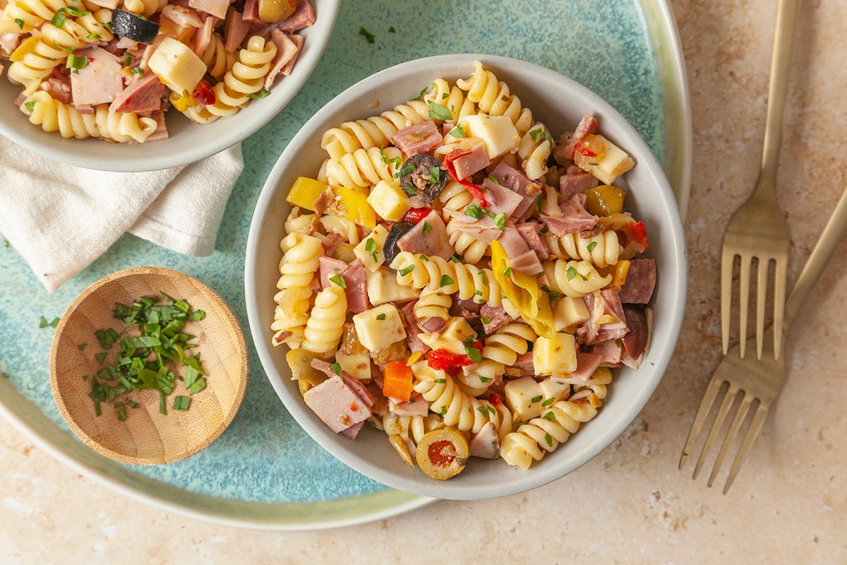 overhead shot of A bowl of rotini pasta salad with cubes of cheese, ham, and colorful bell peppers, garnished with herbs; A small bowl of chopped herbs and a fork are nearby on a blue-green plate