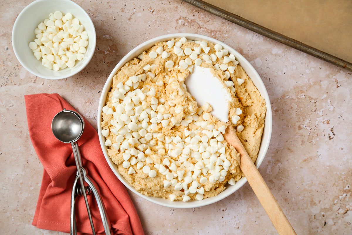 Overhead shot of a large bowl cream butter and sugars until light and fluffy 5-7 minutes; Beat in eggs, orange zest and extracts; In another bowl whisk flour, pudding mix and baking soda; gradually beat into creamed mixture; Stir in white baking chips; spatula; a scoop spoon over a orange napkin; all set on a texture marble surface;