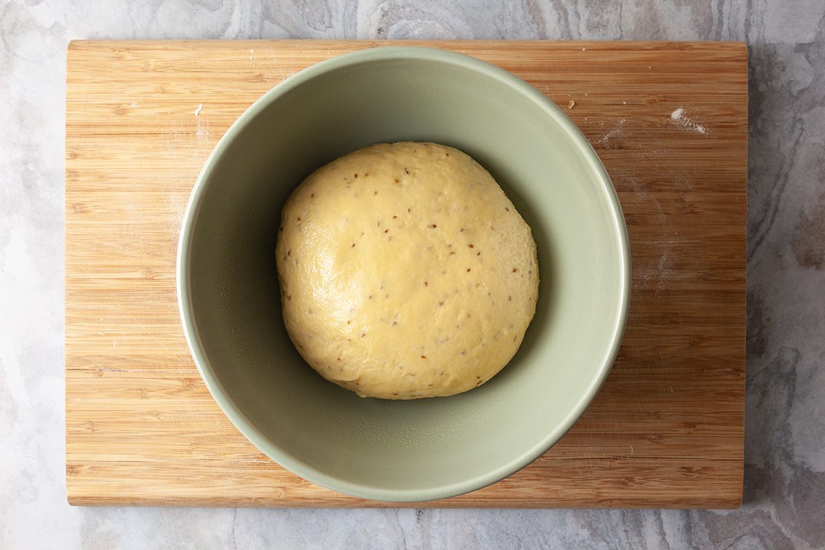 A ball of dough rests in a light green bowl placed on a wooden cutting board, viewed from above.