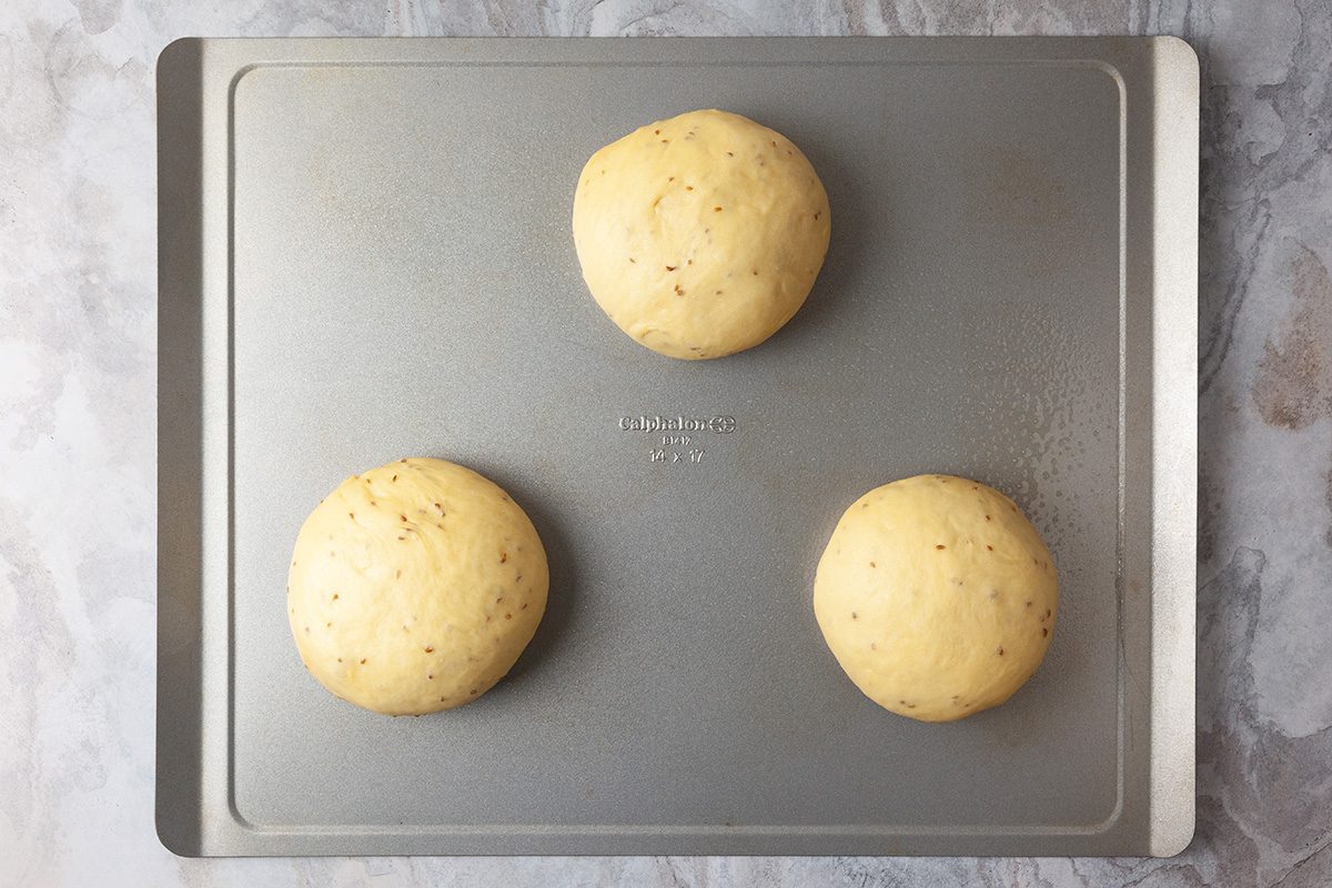 A baking sheet with three evenly spaced, round balls of bread dough resting on it, ready for baking. The dough appears smooth with small specks throughout.