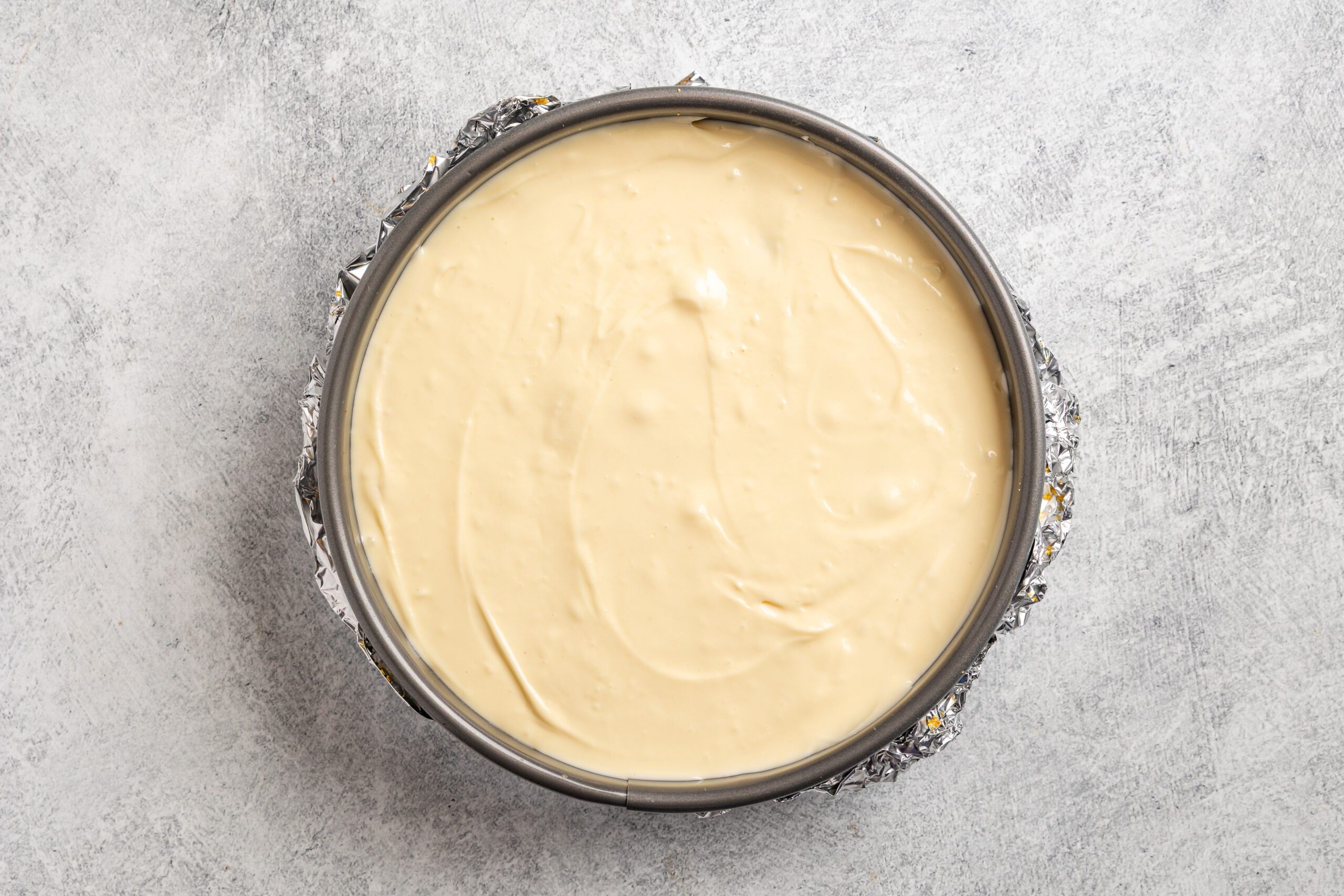 A round springform pan filled with smooth, unbaked cheesecake batter sits on a light gray textured surface, with foil wrapped around the pan's edges.