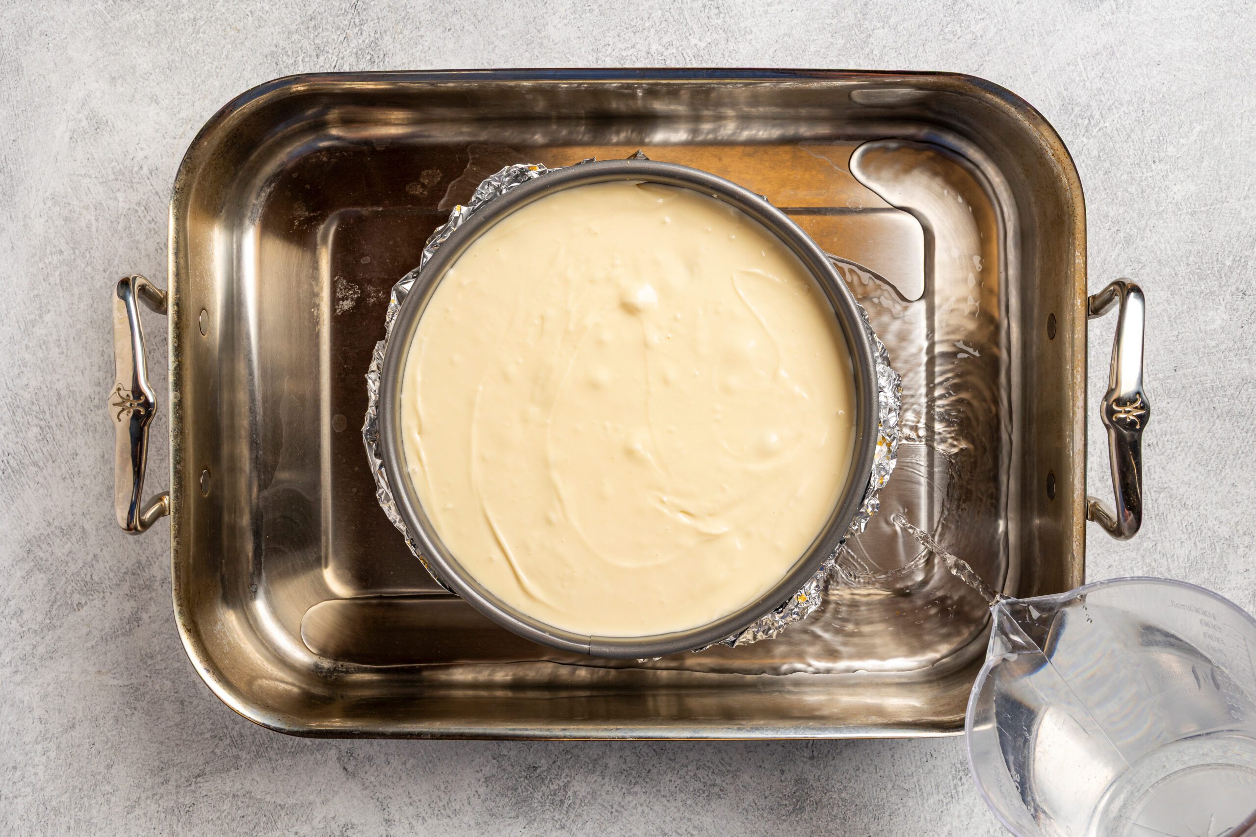 A round springform pan filled with cheesecake batter sits in a large roasting pan, wrapped in foil and ready for a water bath, with a clear pitcher of water nearby on a gray countertop.