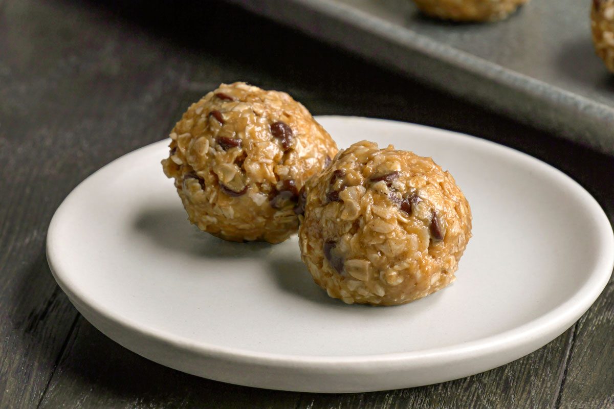 Close-up shot of two peanut butter oatmeal balls with chocolate chips, placed on a small white plate atop a dark wooden surface; A baking tray with more oatmeal balls is visible in the background;