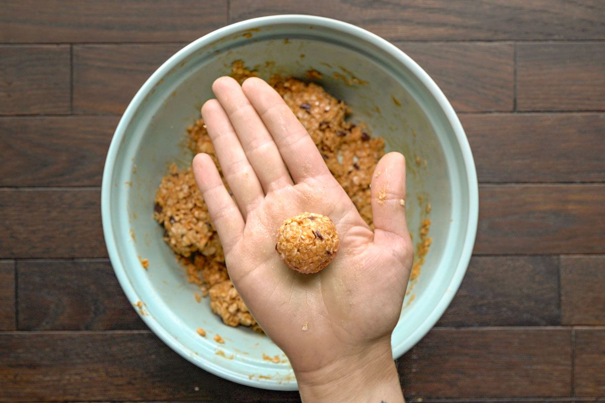 Overhead shot of a hand holding a small; round ball of unbaked cookie dough above a bowl filled with more dough; set on a wooden surface;