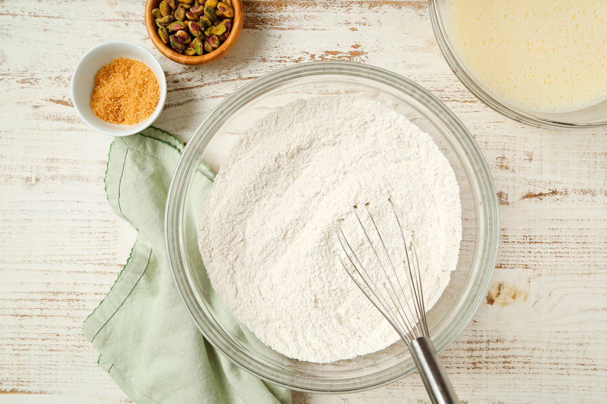overhead shot of a glass bowl filled with flour and a whisk sits on a white wooden surface, Nearby are a green cloth, a small bowl of brown sugar, a bowl of pistachios, and a bowl of liquid batter