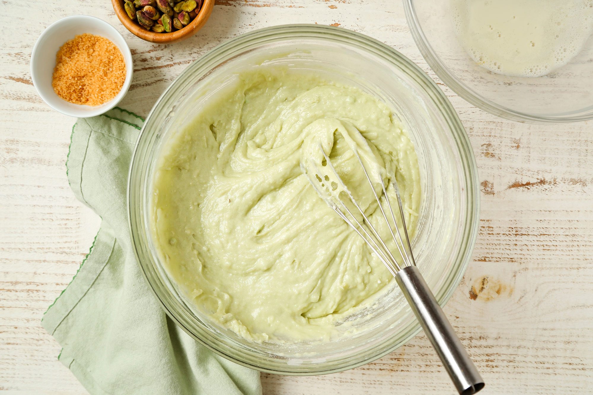 overhead shot of a glass bowl with pale green batter and a whisk, on a light wooden surface, Nearby are a green cloth, a small bowl of brown sugar, a bowl of pistachios, and part of a glass bowl with liquid