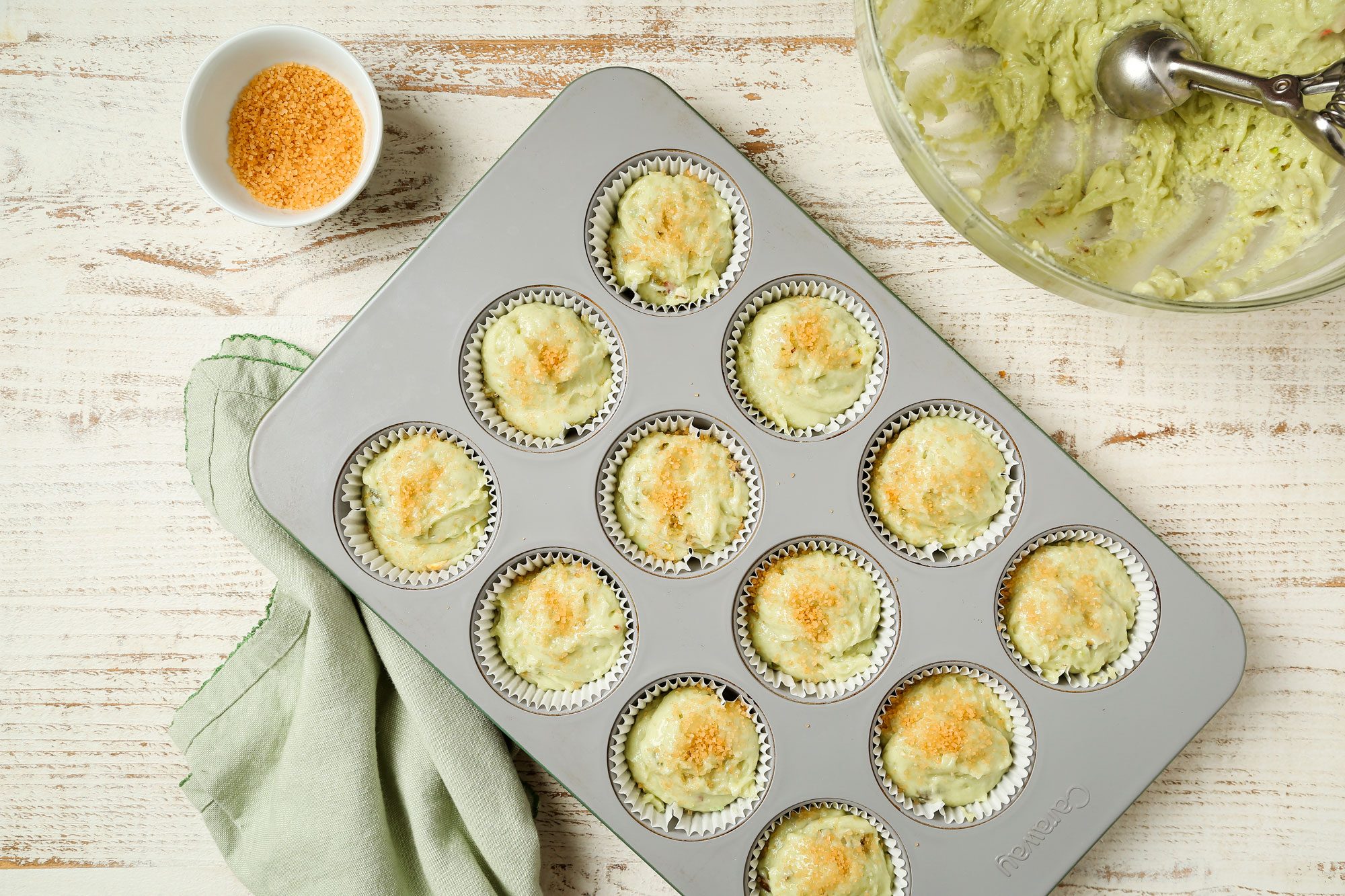 overhead shot of a muffin tin with twelve green, baked treats in paper liners sits on a light wooden surface, Nearby are a green cloth, a bowl of green batter, and a small bowl of breadcrumbs