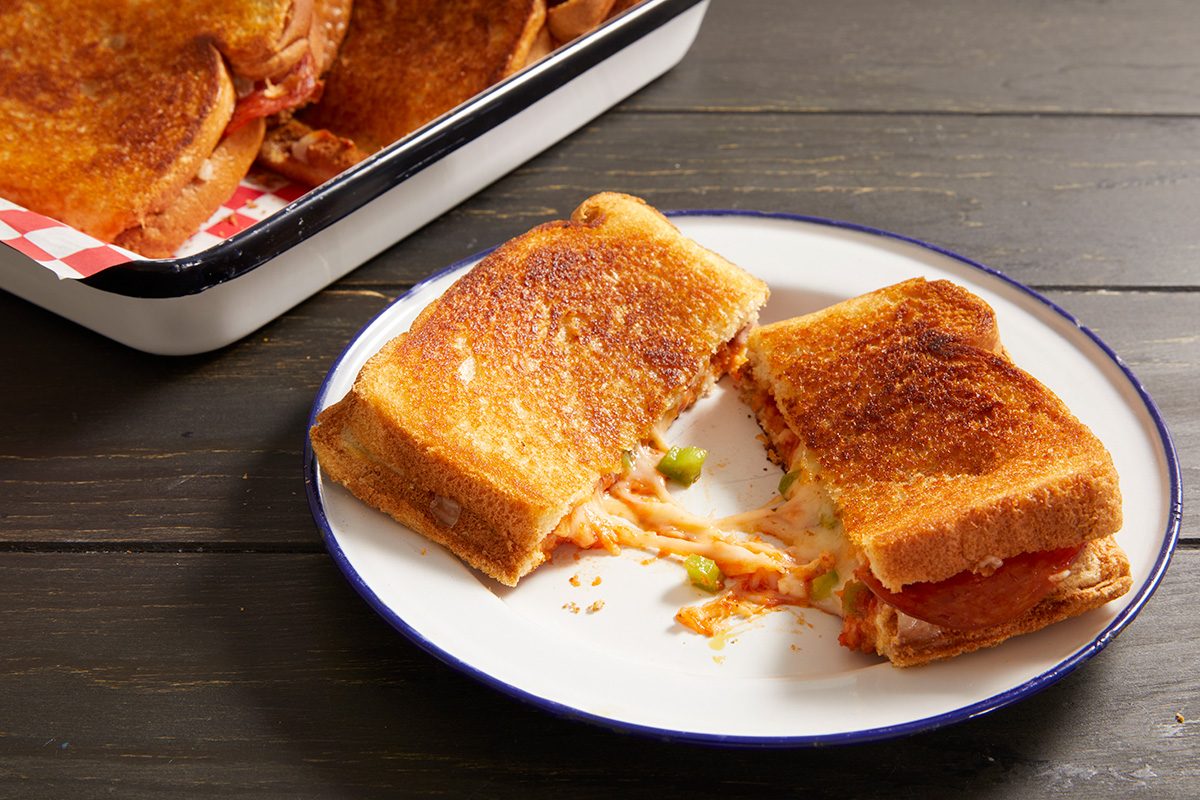 A grilled sandwich cut in half on a white plate, revealing melted cheese and vegetables inside. A tray with more sandwiches is in the background on a dark wooden table.