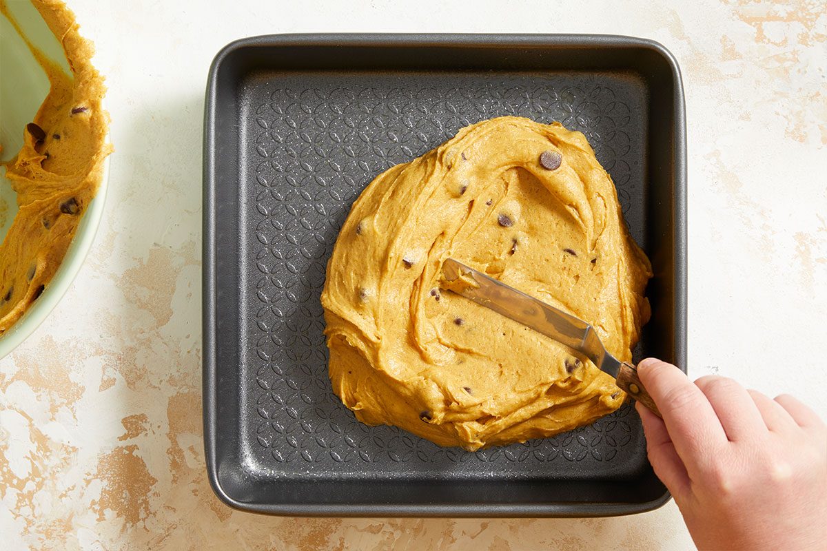 A hand uses a spatula to spread chocolate chip cookie dough evenly in a square baking pan. Some dough is visible on the side of a nearby mixing bowl.